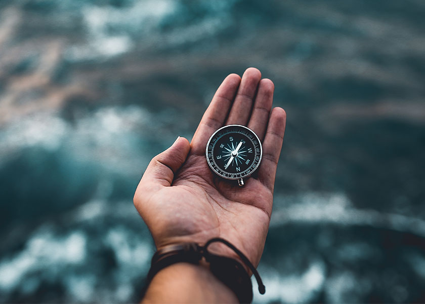 A man's hand holding a compass, extended over ocean waters.